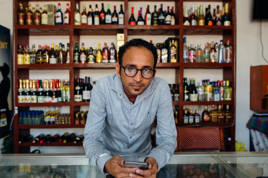 Store clerk leaning on counter waiting to help customer in liquor store. Rows of bottles behind him.